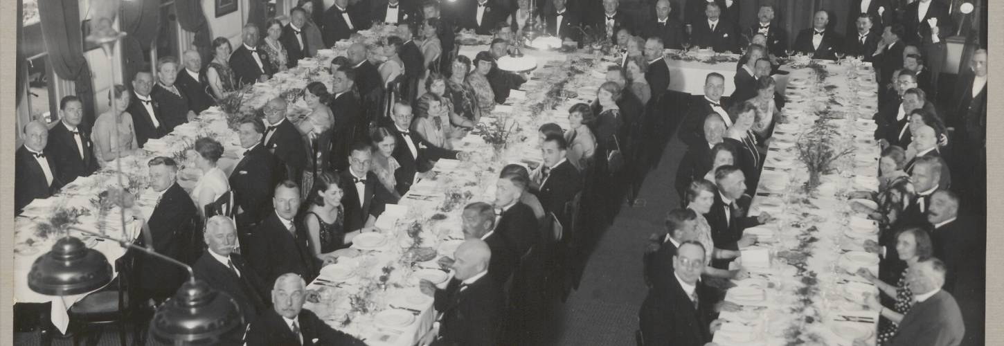 zwart wit foto van een grote groep netje geklede mensen aan lange eettafels