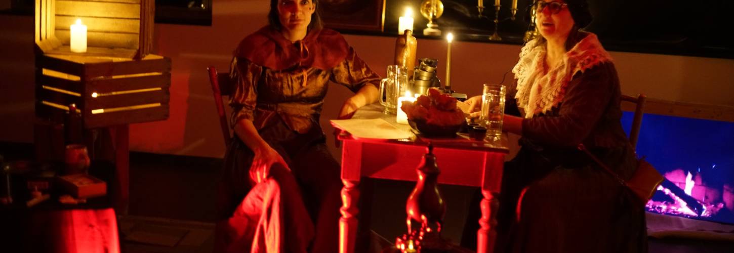 twee vrouwen in een ouderwetse herberg zittend aan tafel met kaarslicht en een haard
