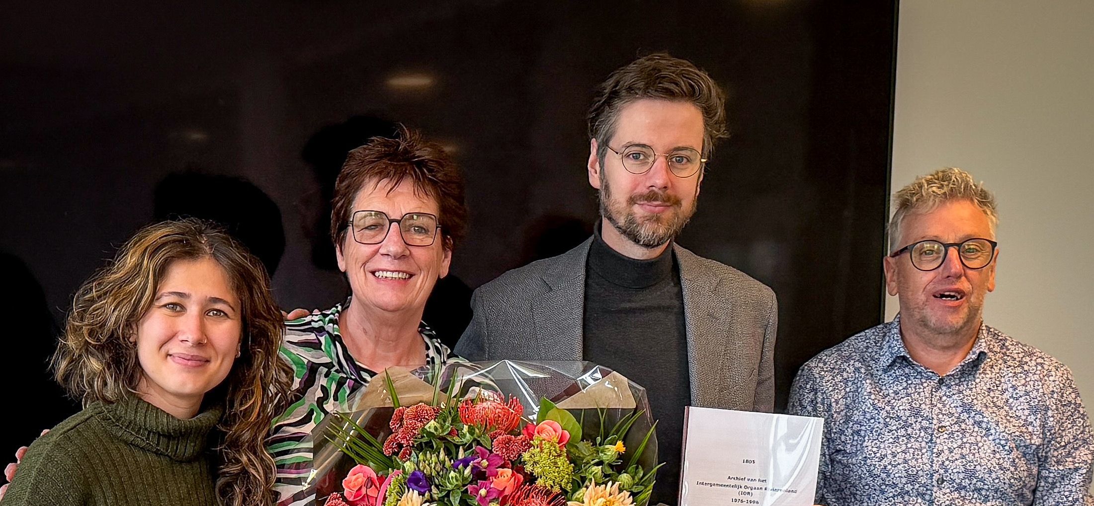 2 vrouwen en 2 mannen, 1 vrouw houdt een bos bloemen vast, 1 man houdt een rapport omhoog