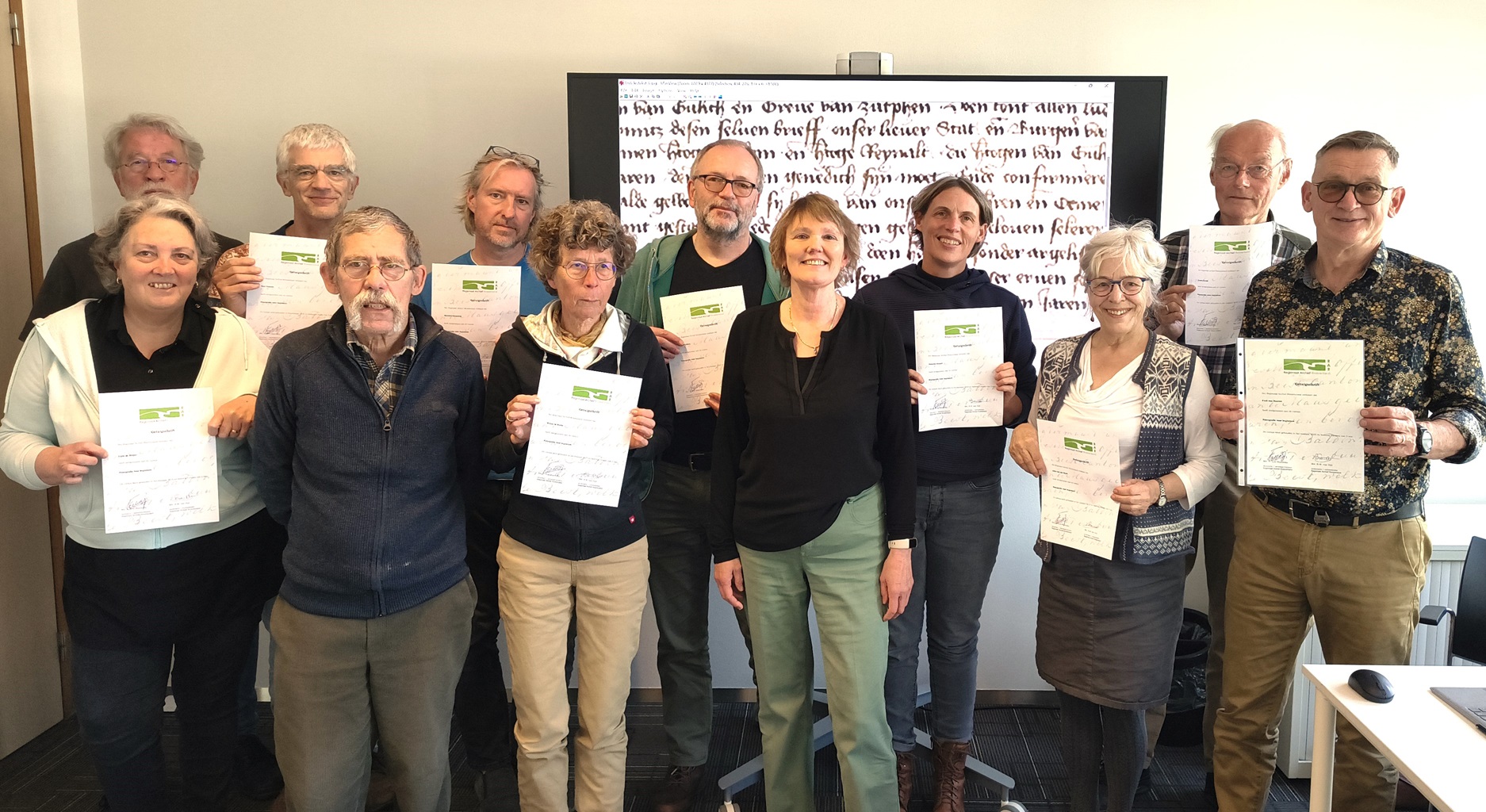 een groep mensen houdt een certificaat omhoog met op de achtergrond een scherm met oud schrift